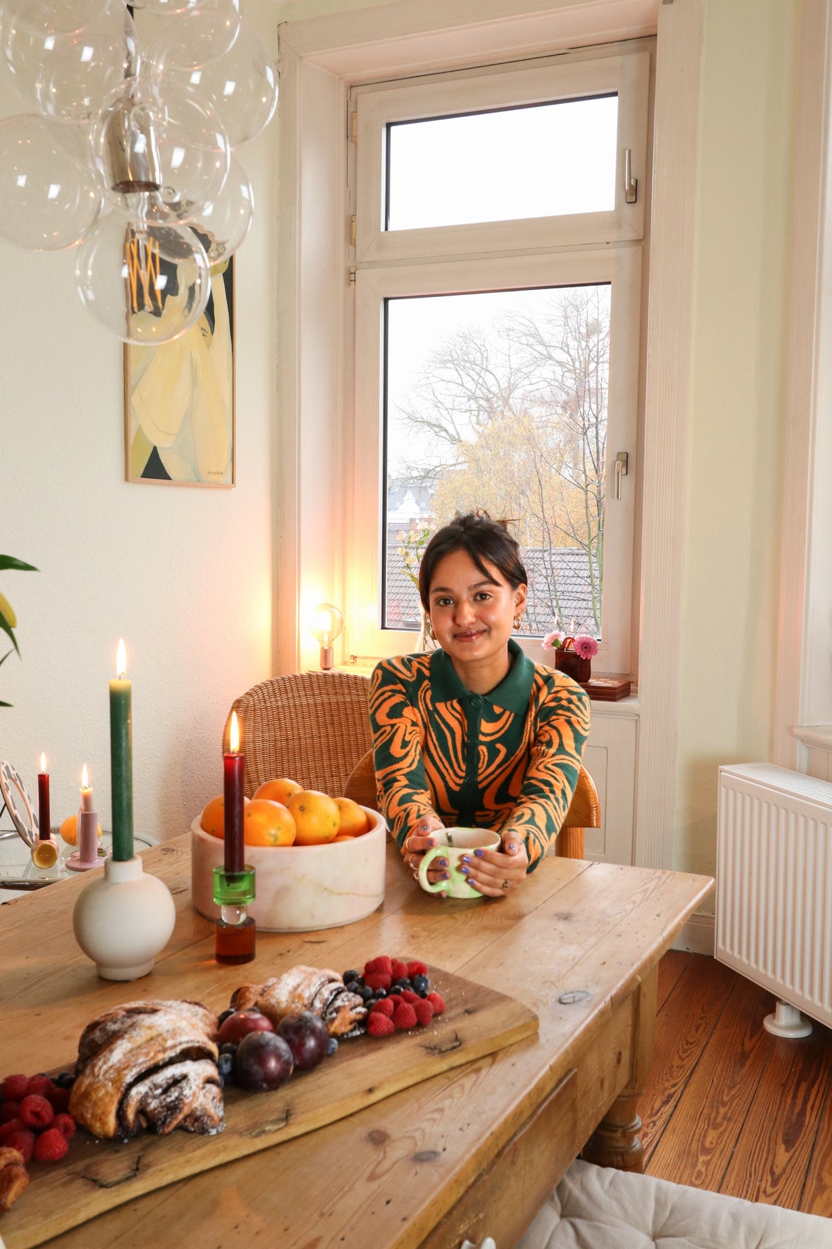 A young woman sits with a mug at a wooden table with fruit, pastries, and candles.