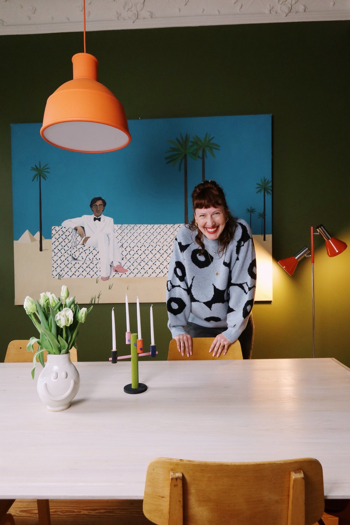 A smiling woman stands by a dining table in front of a large painting and a green wall.