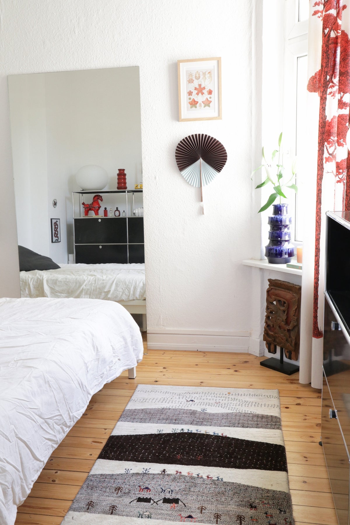 Bedroom with wooden floor, white bed, large mirror, and a patterned rug.