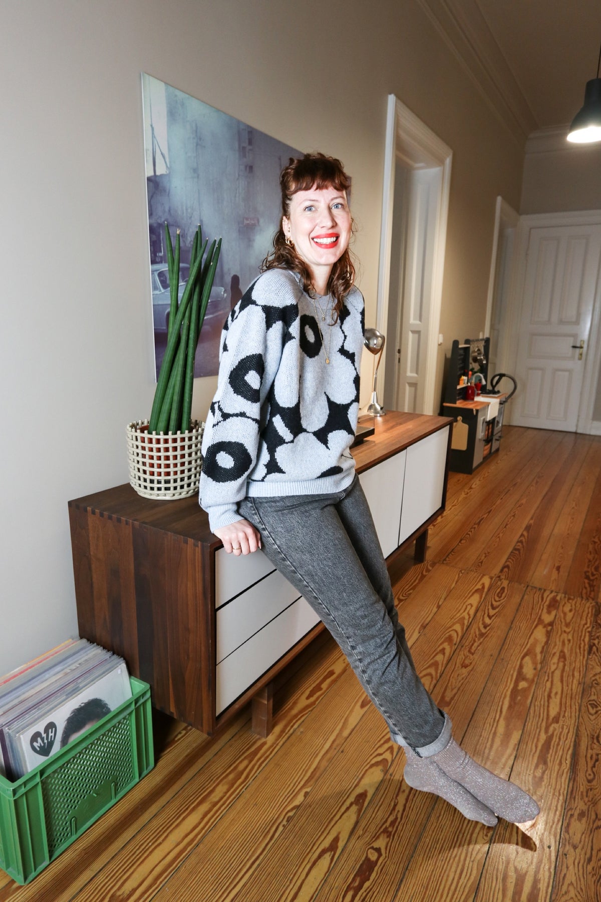 Melanie leans smiling against a wooden sideboard in a hallway with wooden floors.