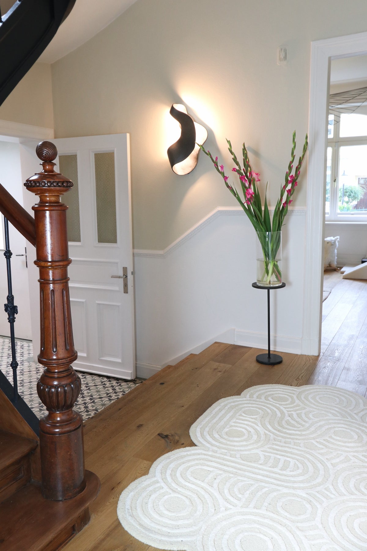 Bright hallway with wooden stairs, wall lamp, vase of flowers, and a light rug on parquet floor.