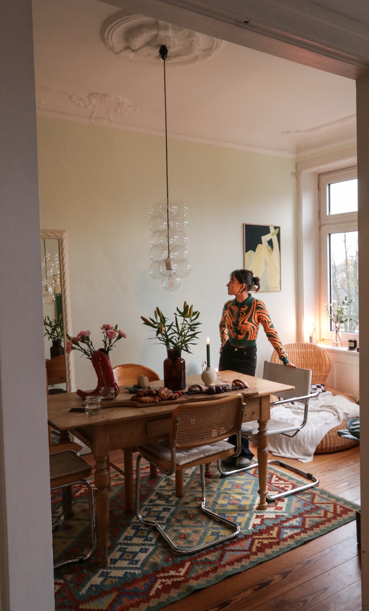 A woman stands in a dining room by a wooden table with chairs on a patterned rug.