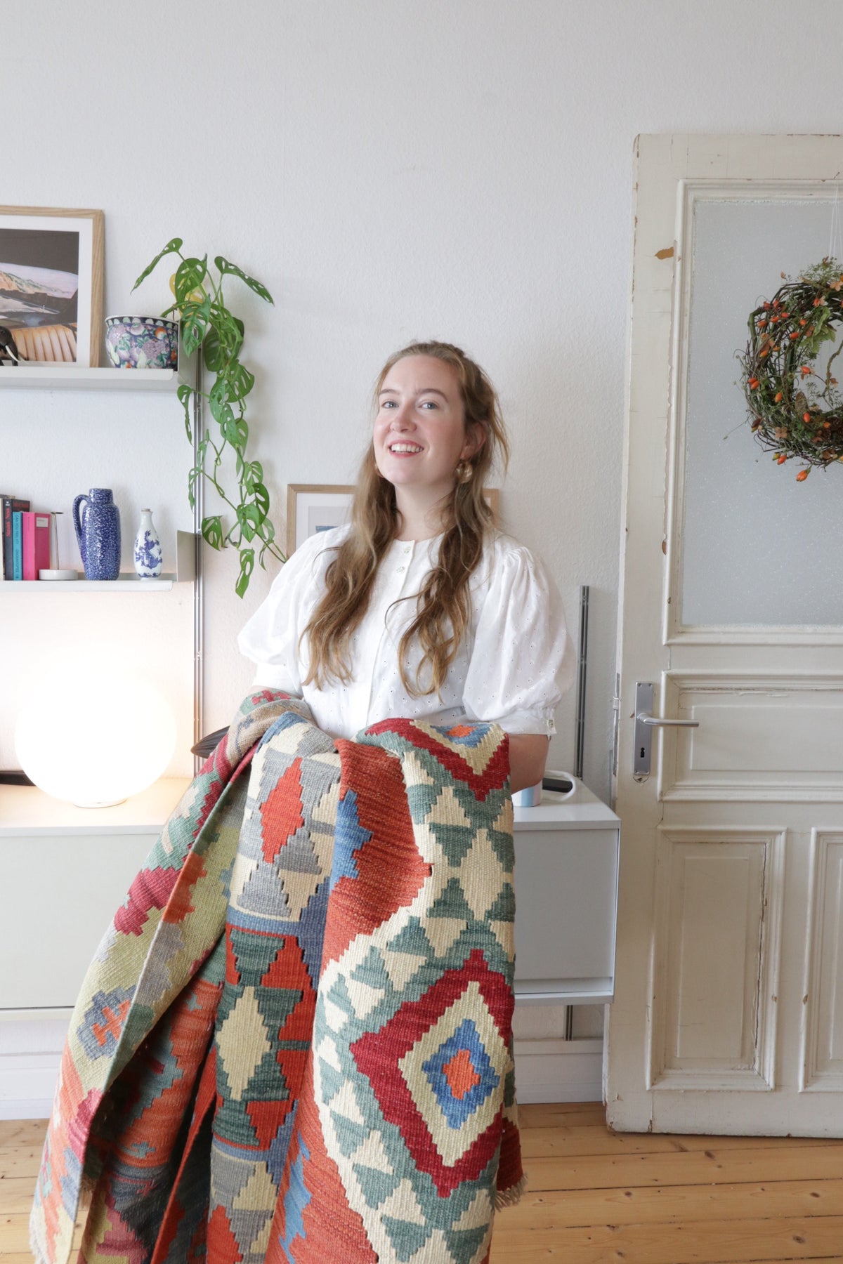 A smiling woman holds a colorful rug with geometric patterns in a bright room.