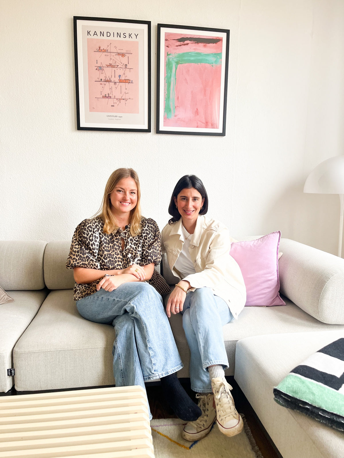 Two women sitting on a sofa in a living room with art prints on the wall and a rug on the floor.