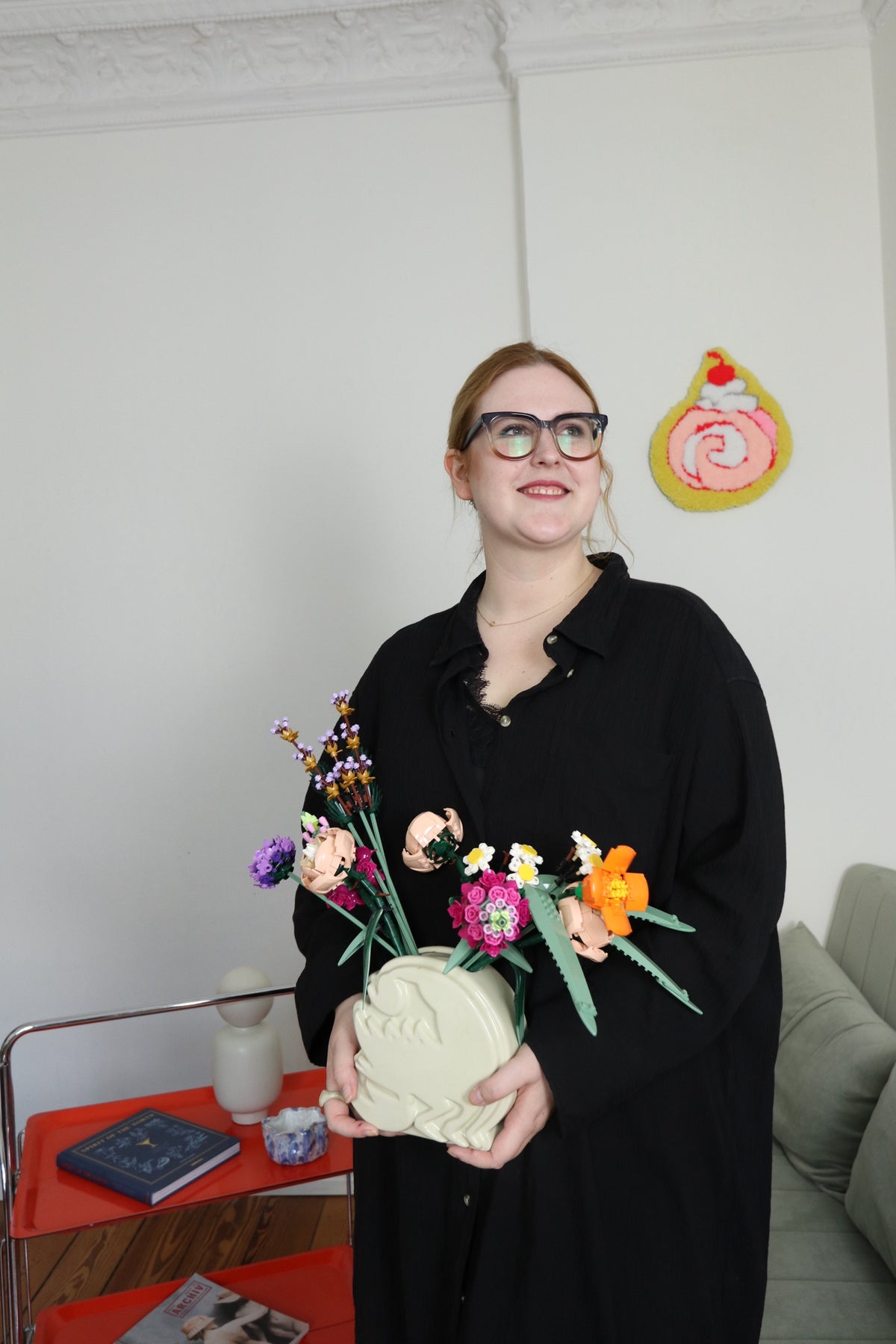 Woman with glasses in a living room holding a vase of colorful toy block flowers.