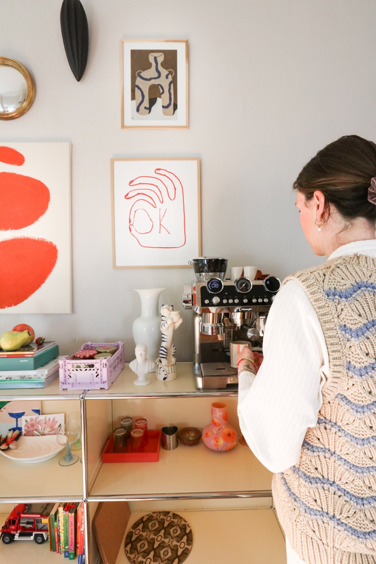A woman at an espresso machine on a shelf unit, with framed art on the wall above.