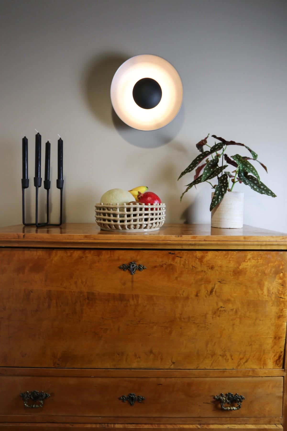 Wooden chest of drawers with a fruit bowl, candle holder, plant, and wall light.