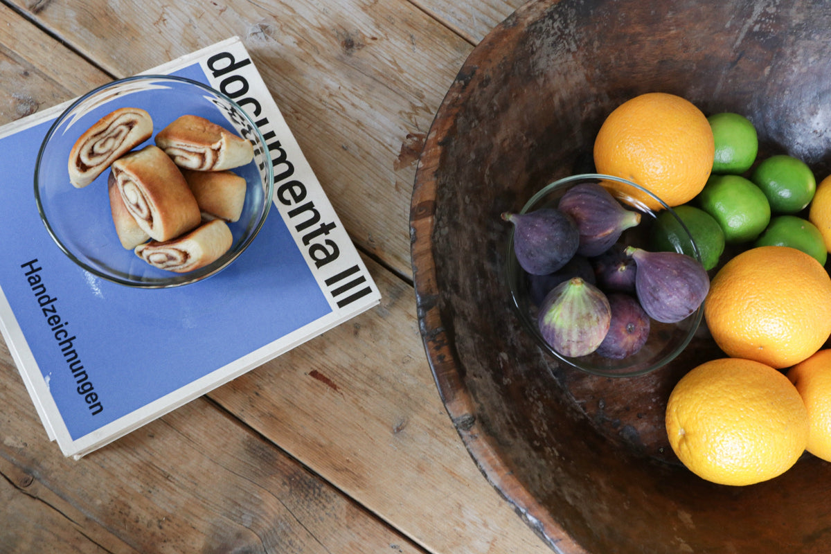Wooden table with a book, a bowl of pastries, and a wooden bowl with figs, oranges, and limes.