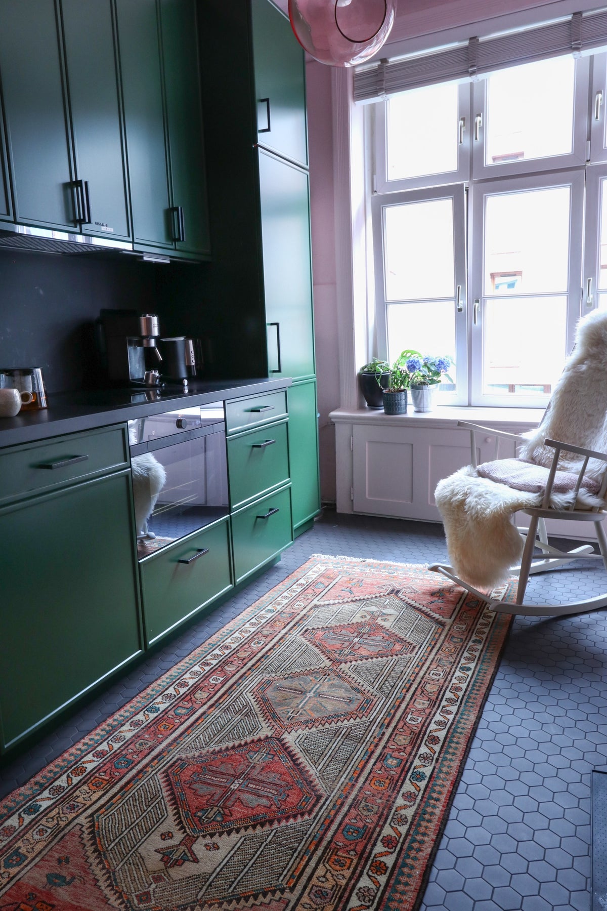 Green kitchen with a patterned rug on a dark tiled floor and a rocking chair by the window.