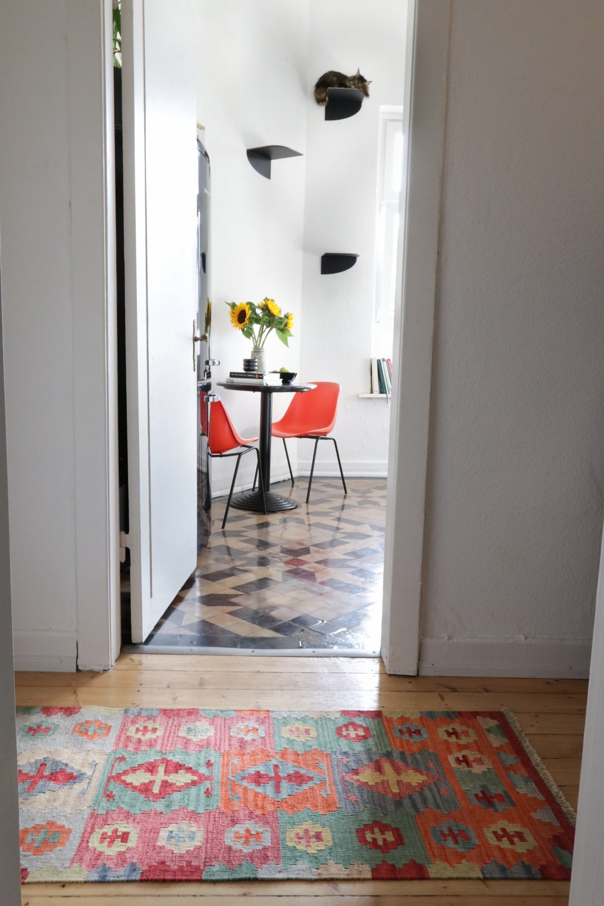 Colorful rug in the hallway facing an open door to a kitchen with table, red chairs, and tiled floor.