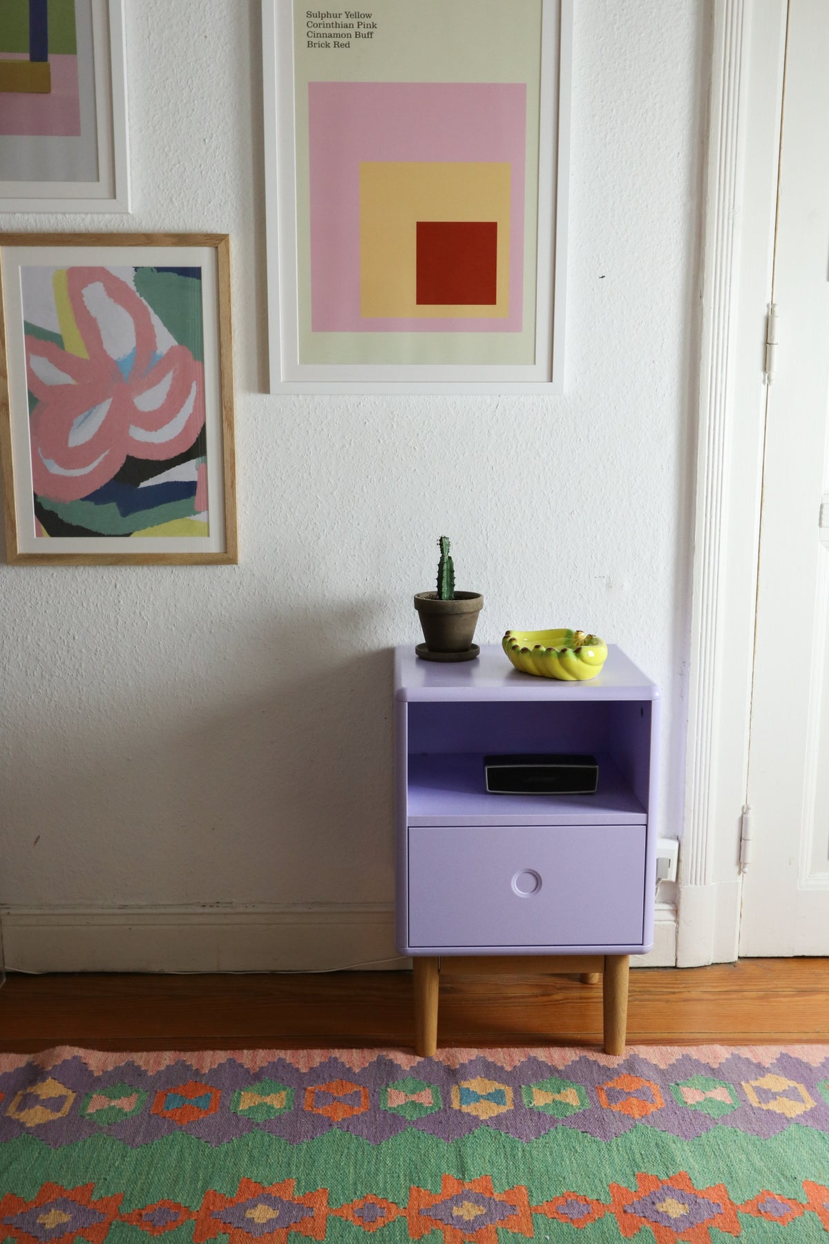 Lilac side table by a white wall with prints, and a colorful rug on the wooden floor.